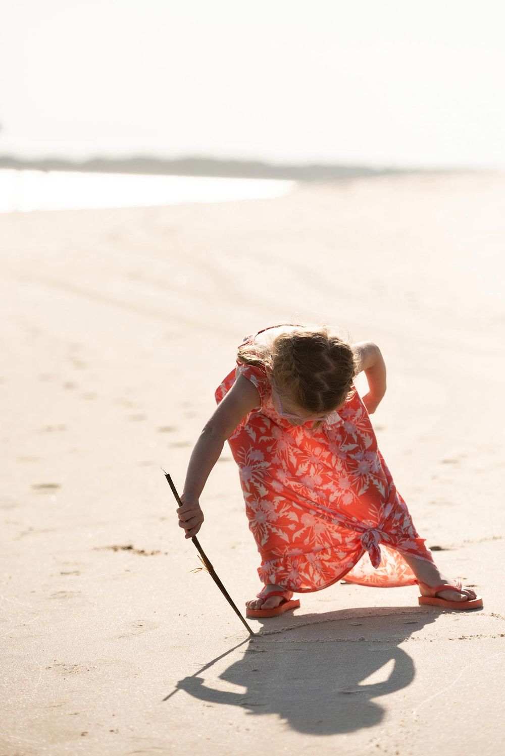Child in orange clothing drawing in the sand at the beach during golden hour.