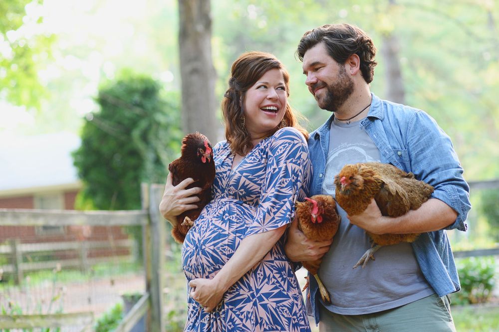 A couple wearing casual clothes holds brown chickens while standing together outdoors in a garden setting.