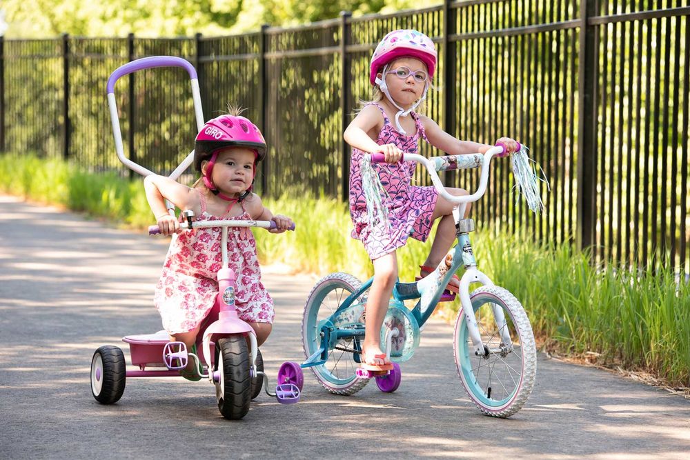 Two children riding bikes on a paved path alongside a black metal fence on a sunny day.