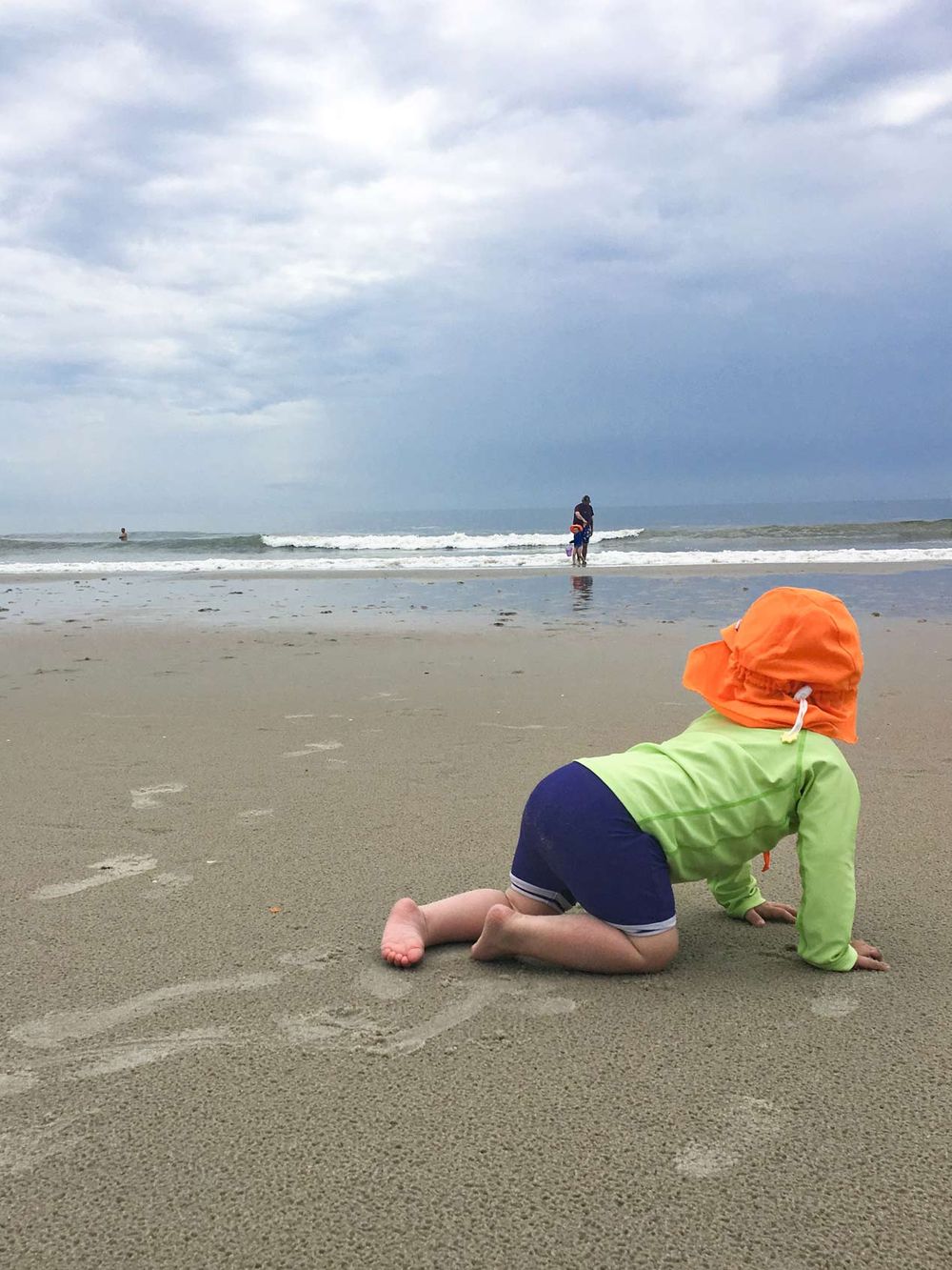 Child in orange hat and colorful clothing crawling on beach sand with ocean waves in background.