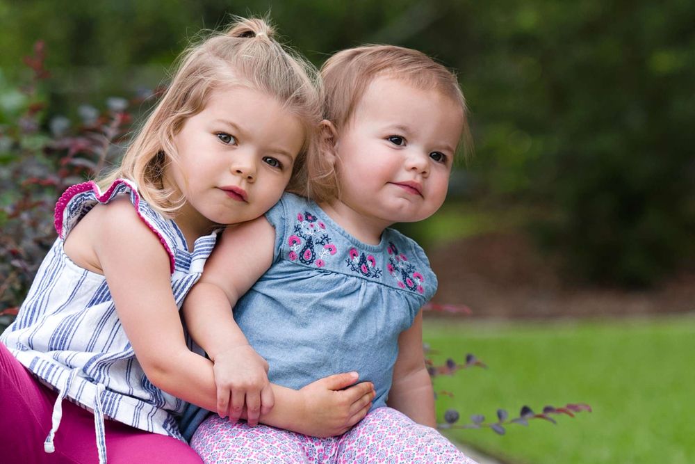 Two young children sitting together outdoors in summer dresses against a garden background.