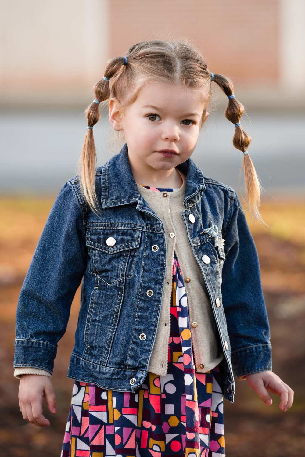 Small child wearing denim jacket and patterned dress stands outdoors in autumn.