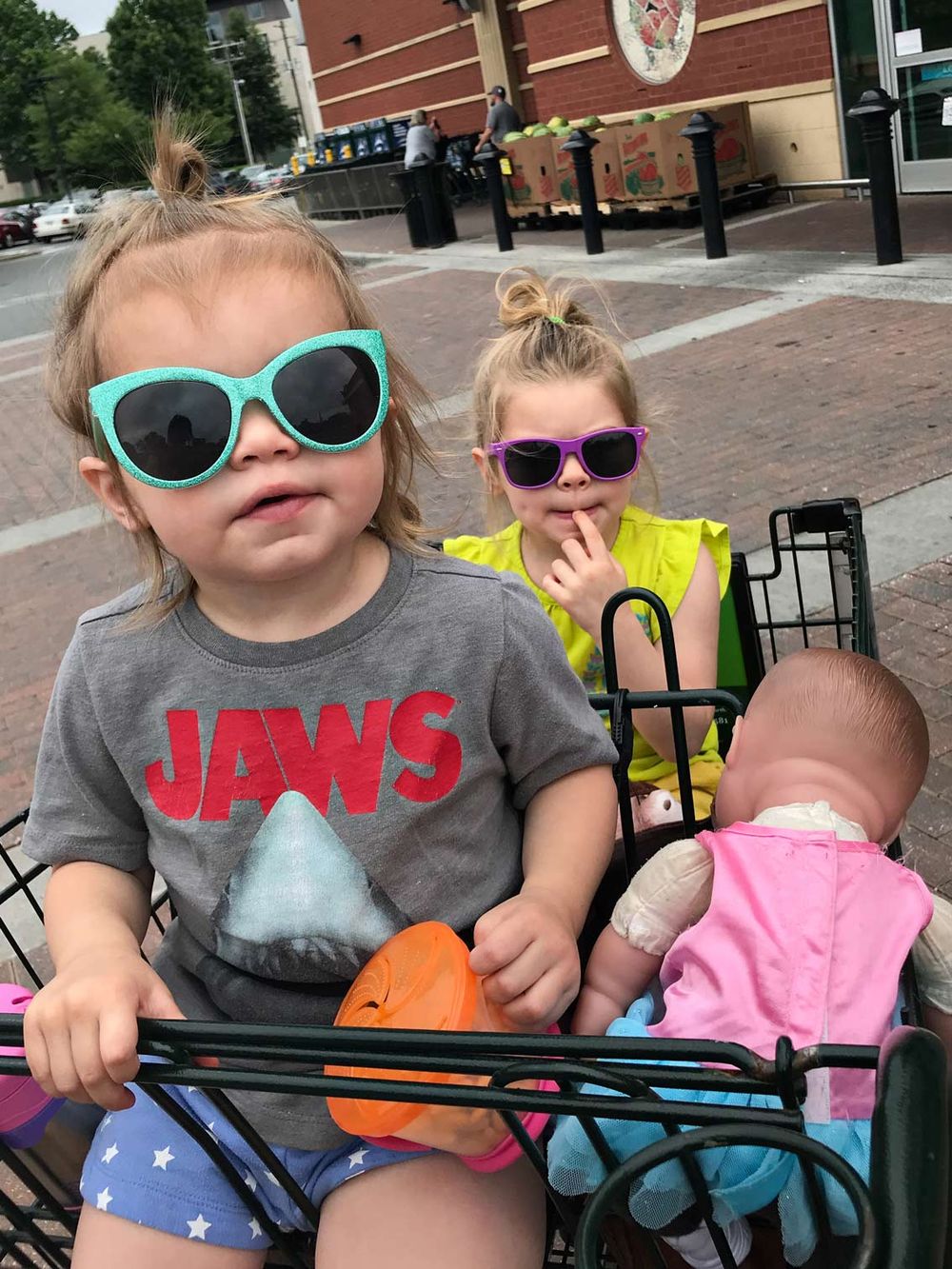 Three children sitting in a shopping cart wearing sunglasses on a brick sidewalk.