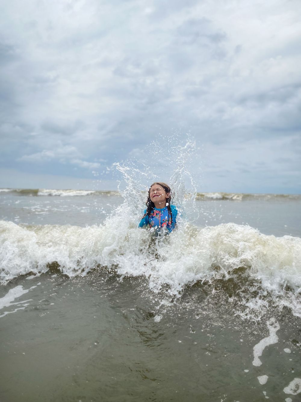 A swimmer enjoying the ocean waves while getting splashed by the surf at the beach on a cloudy day.