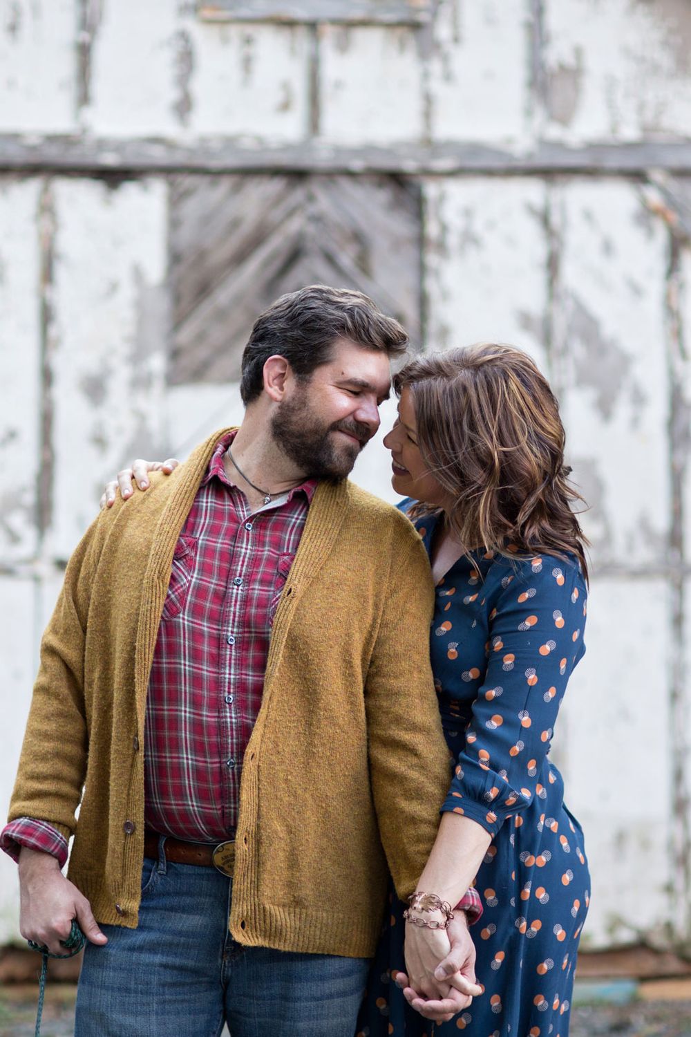 A couple in casual fall attire share an intimate moment while holding hands against a rustic white wall background.