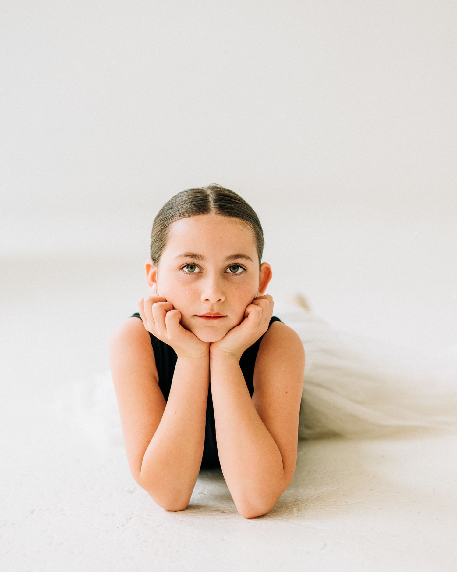 Young ballerina lying on floor with chin resting on hands, wearing black leotard and white tutu.