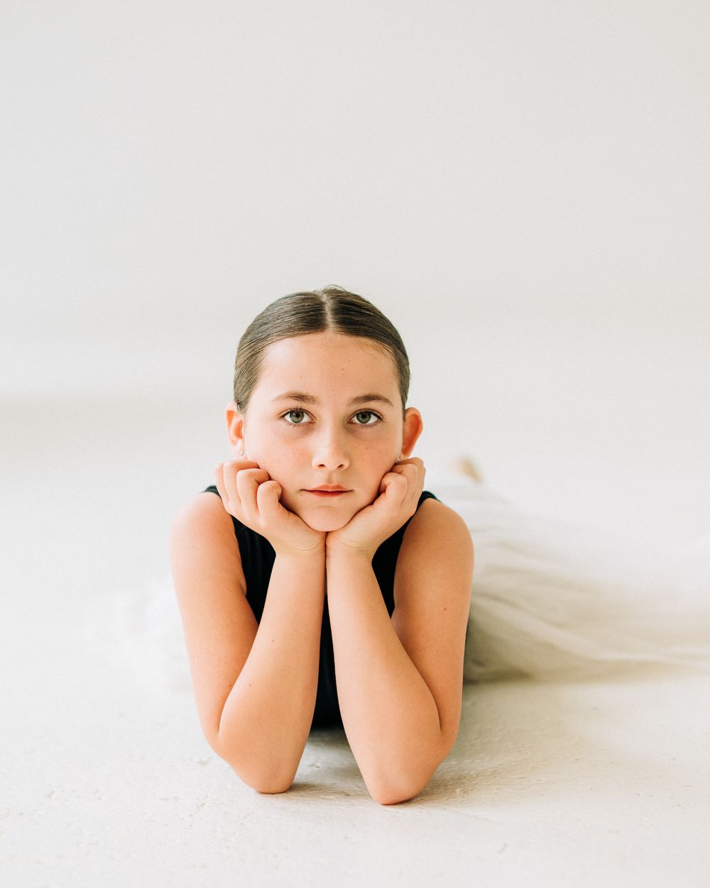 Young ballerina lying on floor with chin resting on hands, wearing black leotard and white tutu.