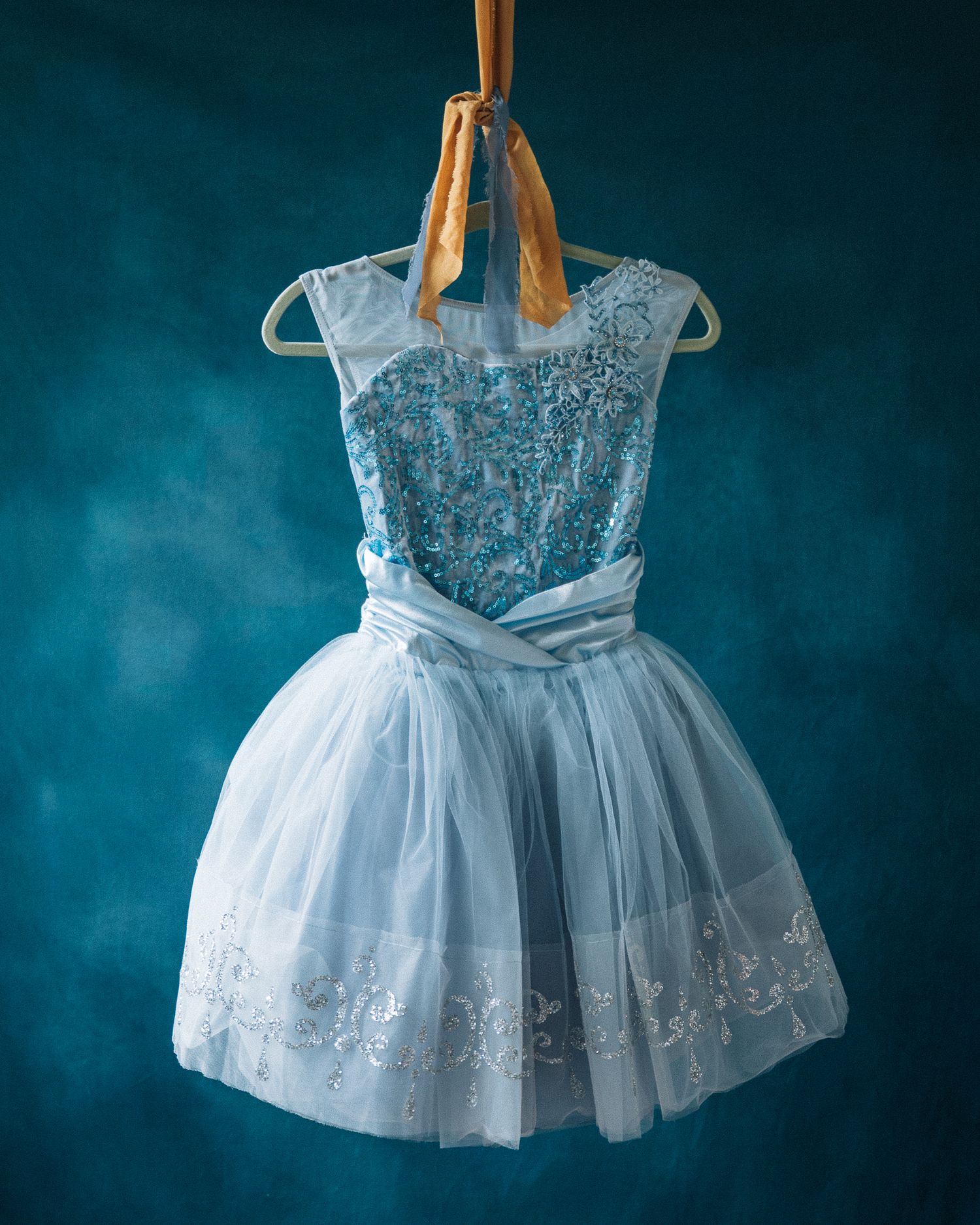Elegant light blue tulle girls dress with lace bodice and satin bow, hanging on a hanger against teal background.