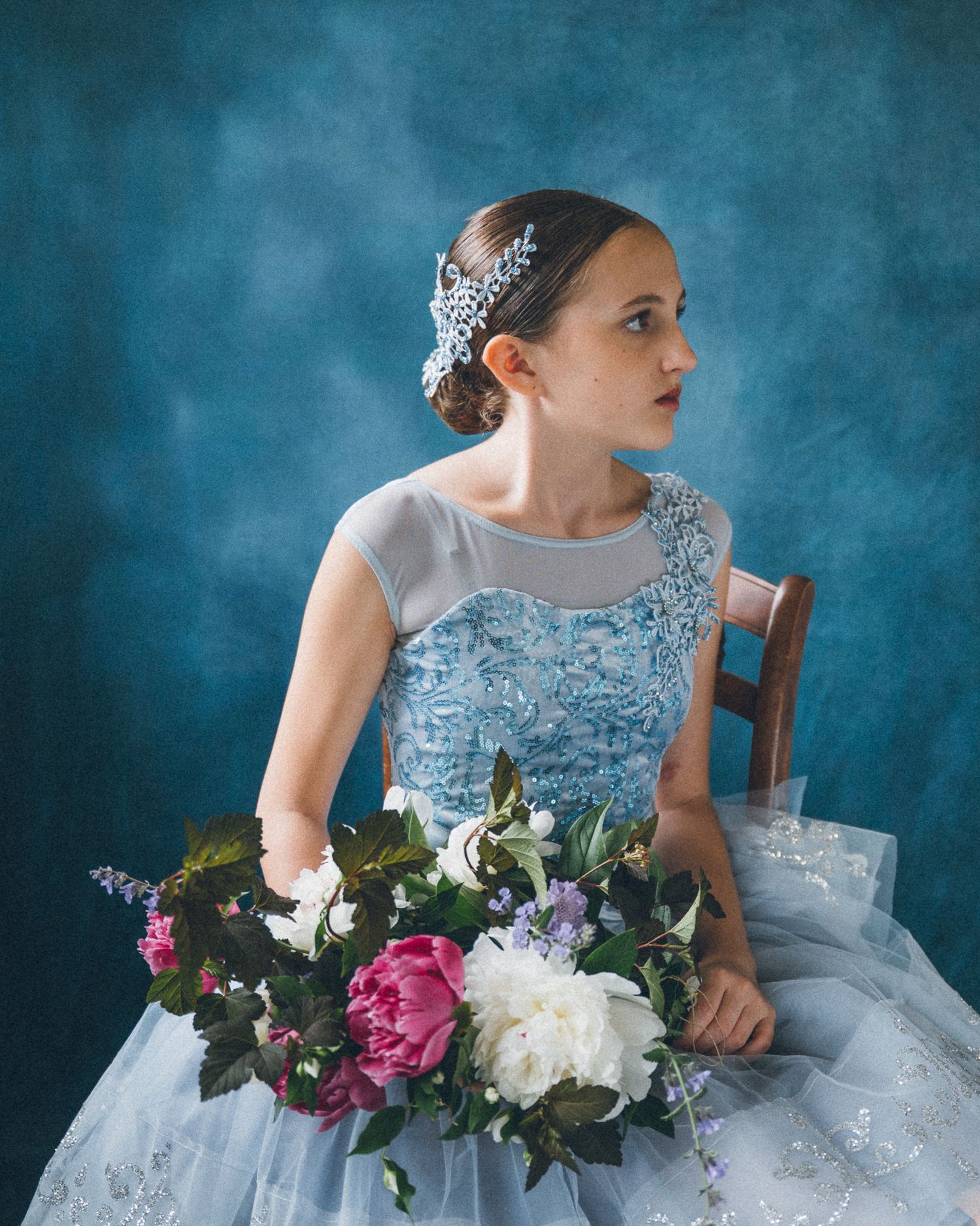 Young girl in blue embroidered gown with floral headpiece holds colorful bouquet while seated against blue backdrop.