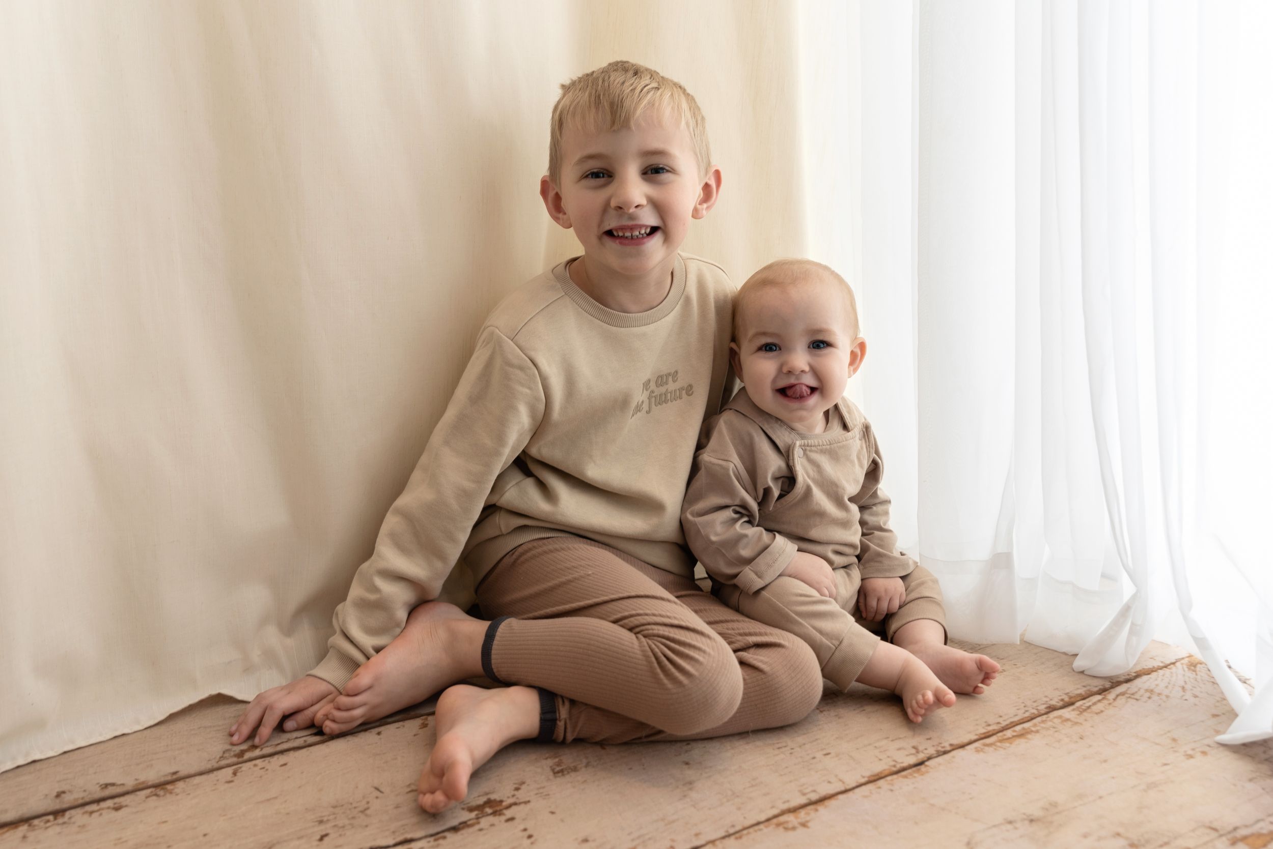 Two young children sit together smiling on a white carpet against cream curtains in natural light.