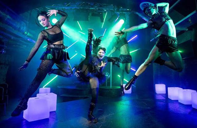 Dancers leap through the air in dramatic poses against neon blue lighting and glowing cube props.