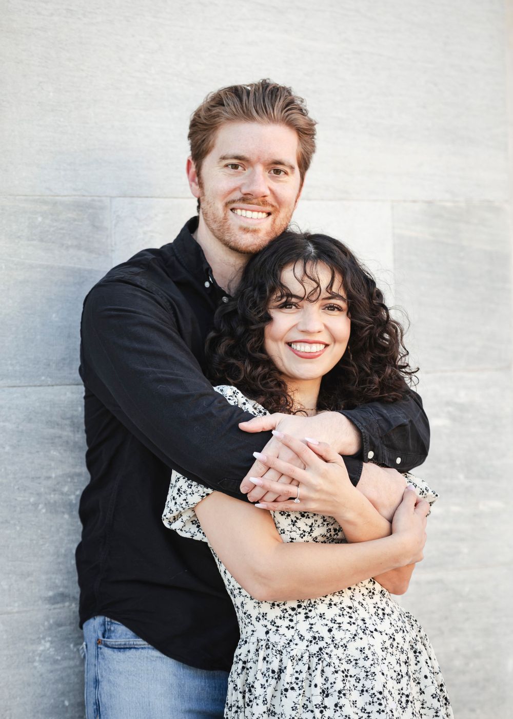 Happy couple embracing and smiling against a light wall while wearing casual black and floral outfits.