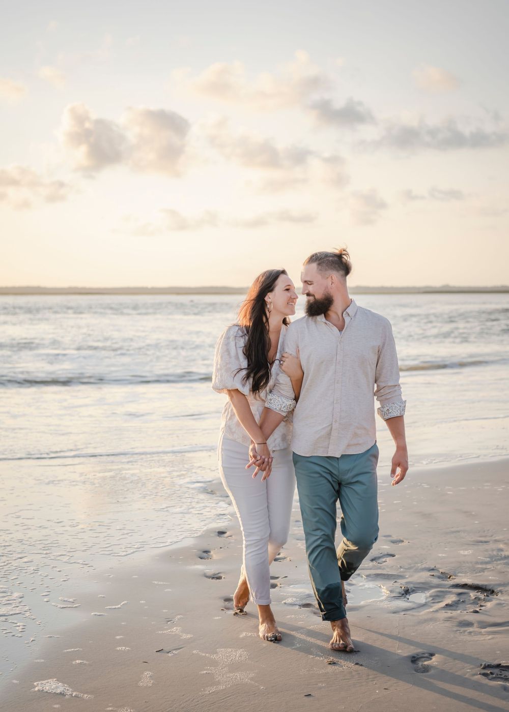A couple walks barefoot together on the beach at sunset holding hands in casual attire.