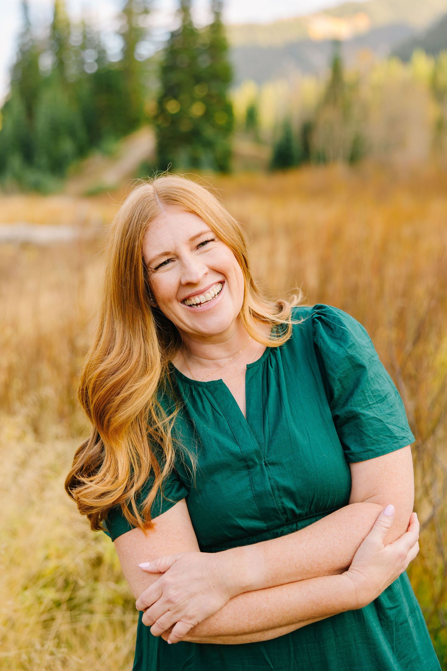 Smiling woman in emerald green dress poses in golden field with mountains in background at sunset.