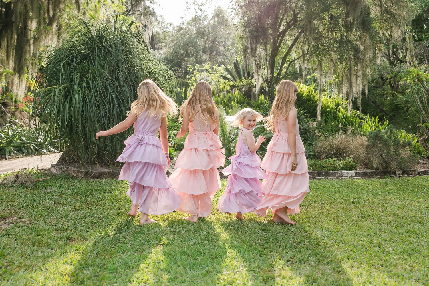 Girls in matching pink dresses sitting together on grass under Spanish moss trees.