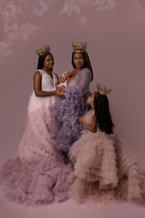 A mother and her three daughters in flowing tulle gowns wearing crowns pose elegantly against mauve backdrop.