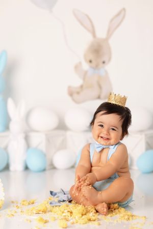 Smiling baby wearing tiny crown sits among blue and white decorations with bunny plush in background.