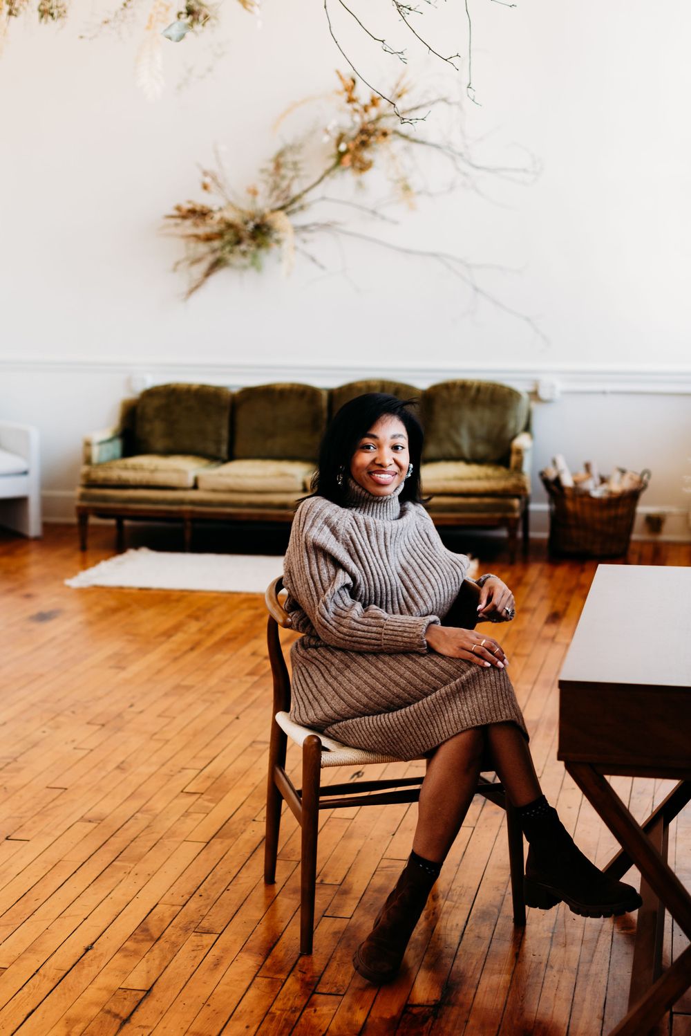 A person in a cozy knit sweater dress sits on a wooden chair in a bright room with hardwood floors and vintage furniture.