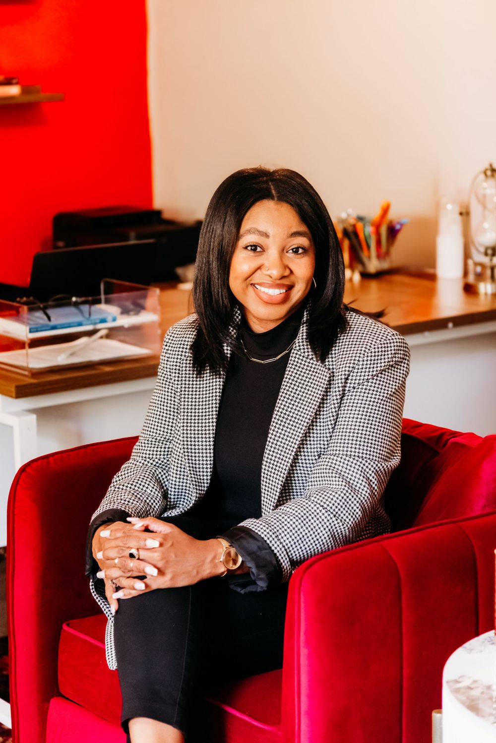 Professional woman wearing a checkered blazer sits confidently in a red armchair against vibrant office decor.