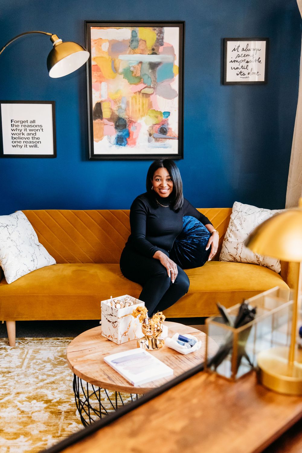 Stylish living room features a mustard yellow couch against deep blue walls with modern abstract art and round coffee table.