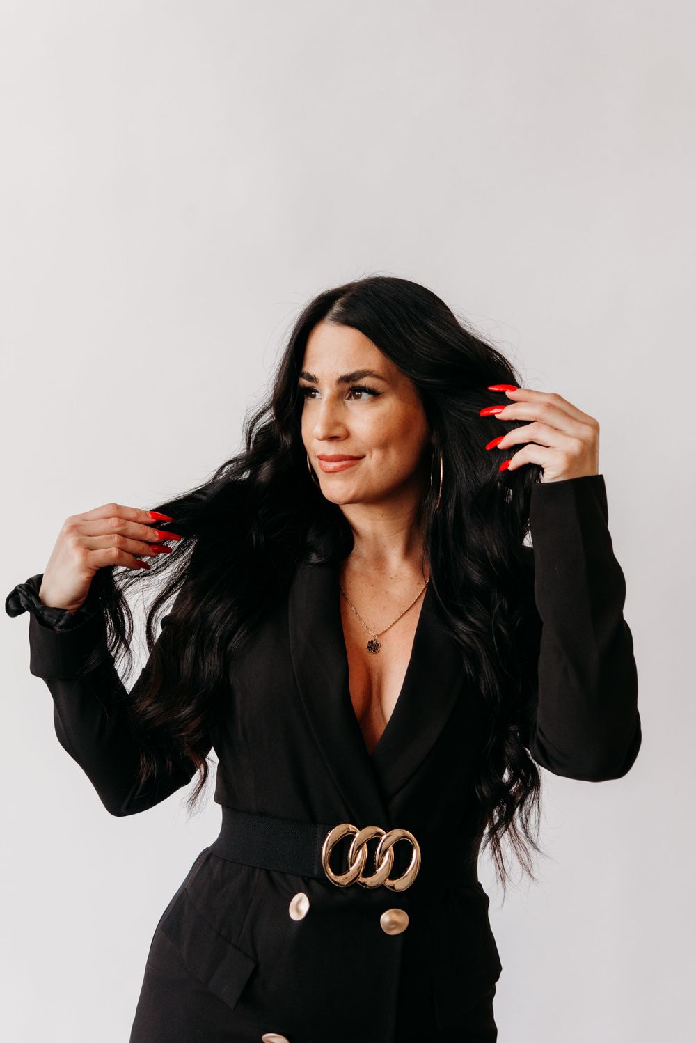 Person wearing a black blazer with gold belt buckle poses against white background with long dark hair and red nails.