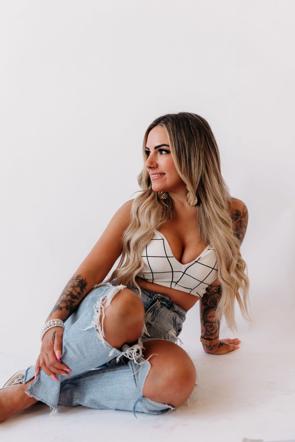 Stylish model in ripped jeans and geometric crop top poses against white backdrop while showcasing tattoo sleeves.
