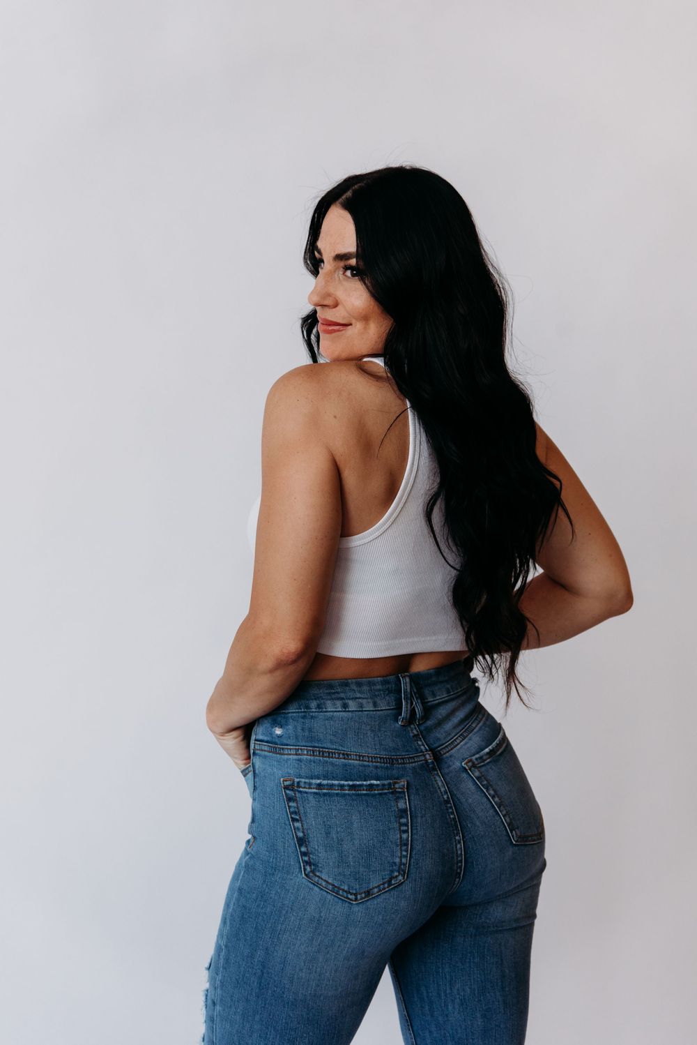 Woman wearing white crop tank top and high waisted blue jeans poses against white wall with back turned to camera.
