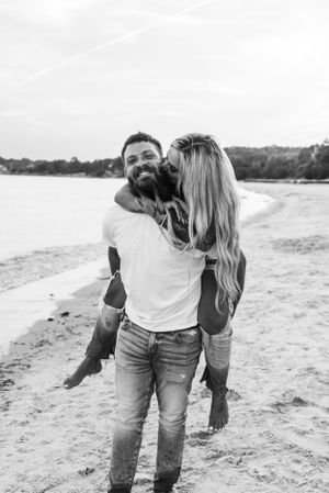 Black and white photo of romantic couple sharing playful moment on sandy beach with water in background.