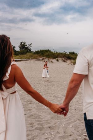 A couple holding hands on a sandy beach with a small figure in a white dress visible in the distance.