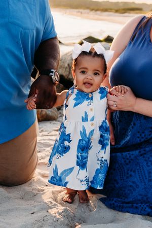 Young child in white and blue floral dress standing on sand at the beach with adults sitting beside them.
