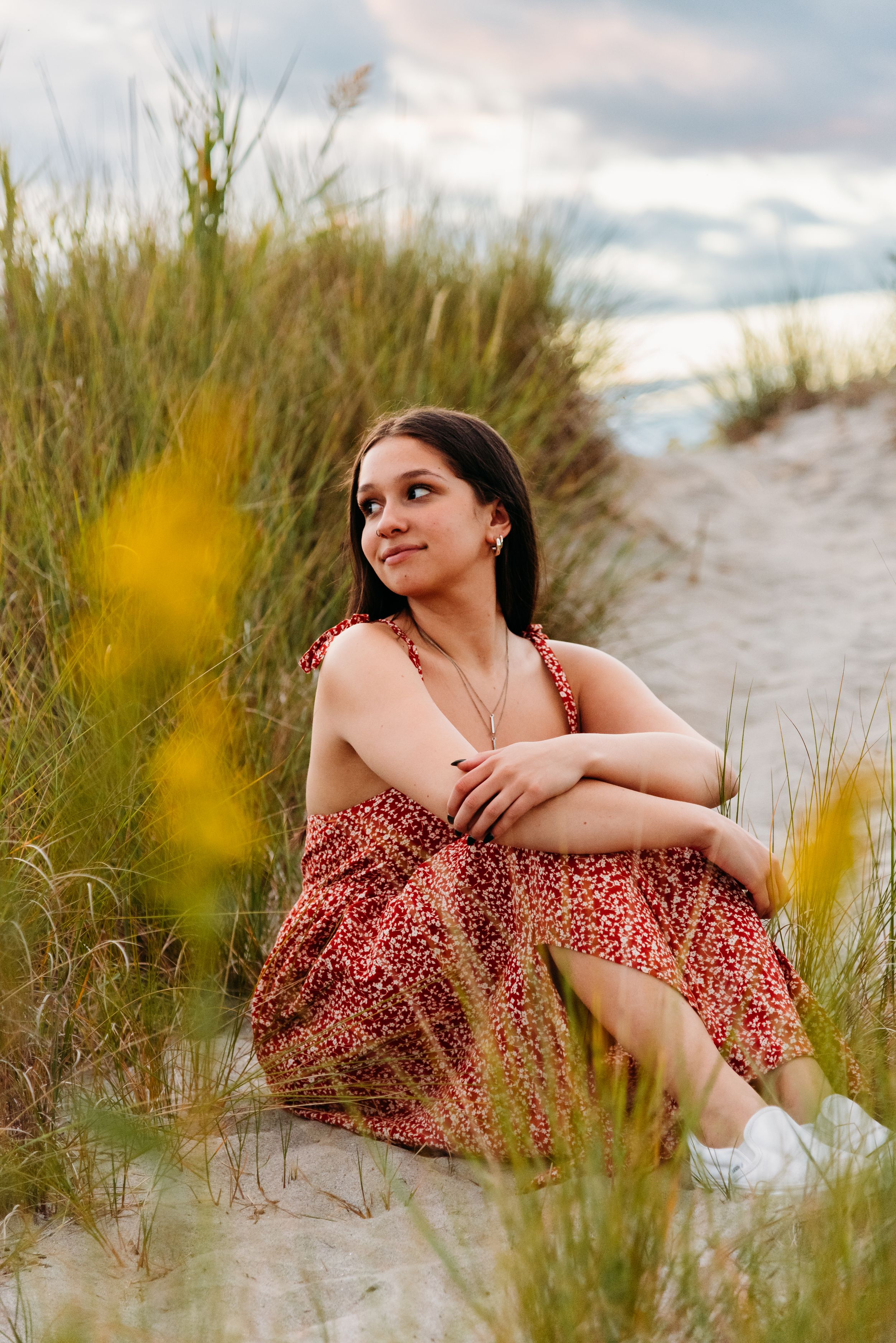 A summer portrait in a floral sundress sitting among beach grass and dunes on a cloudy day.