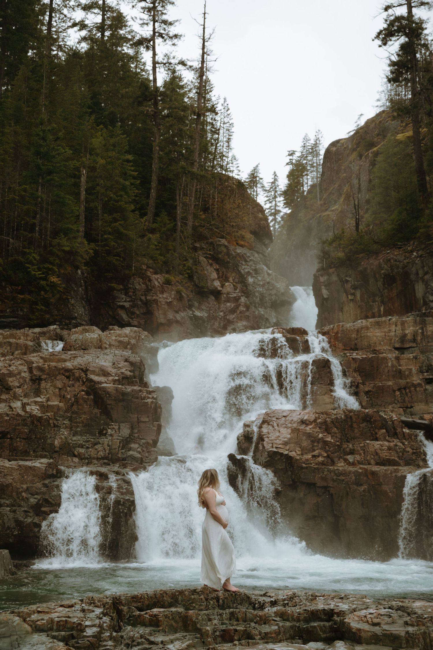 A picturesque waterfall cascades down rocky cliffs in a pine forest while a figure in white stands on the rocks below.