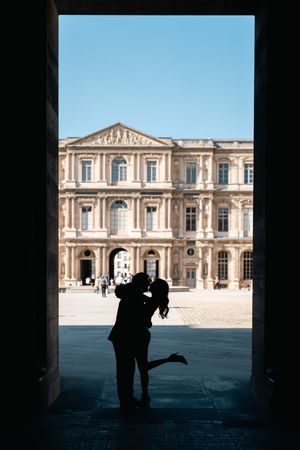 Silhouette d’un couple de mariés sous une arche sombre avec le Louvre, batiment historique illuminé en arrière-plan.