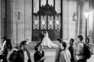 Mariée debout dans une grande porte en bois tandis que la foule passe floue devant elle, composition artistique en noir et blanc.