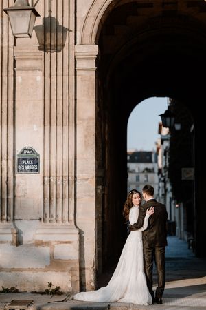 Couple de mariés posant sous une grande arche en pierre dans un décor parisien élégant.