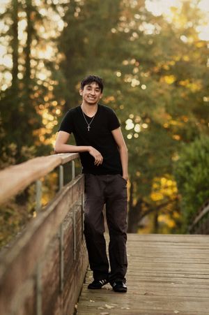 A person in black attire leans casually against a wooden bridge railing during golden hour in a park setting.