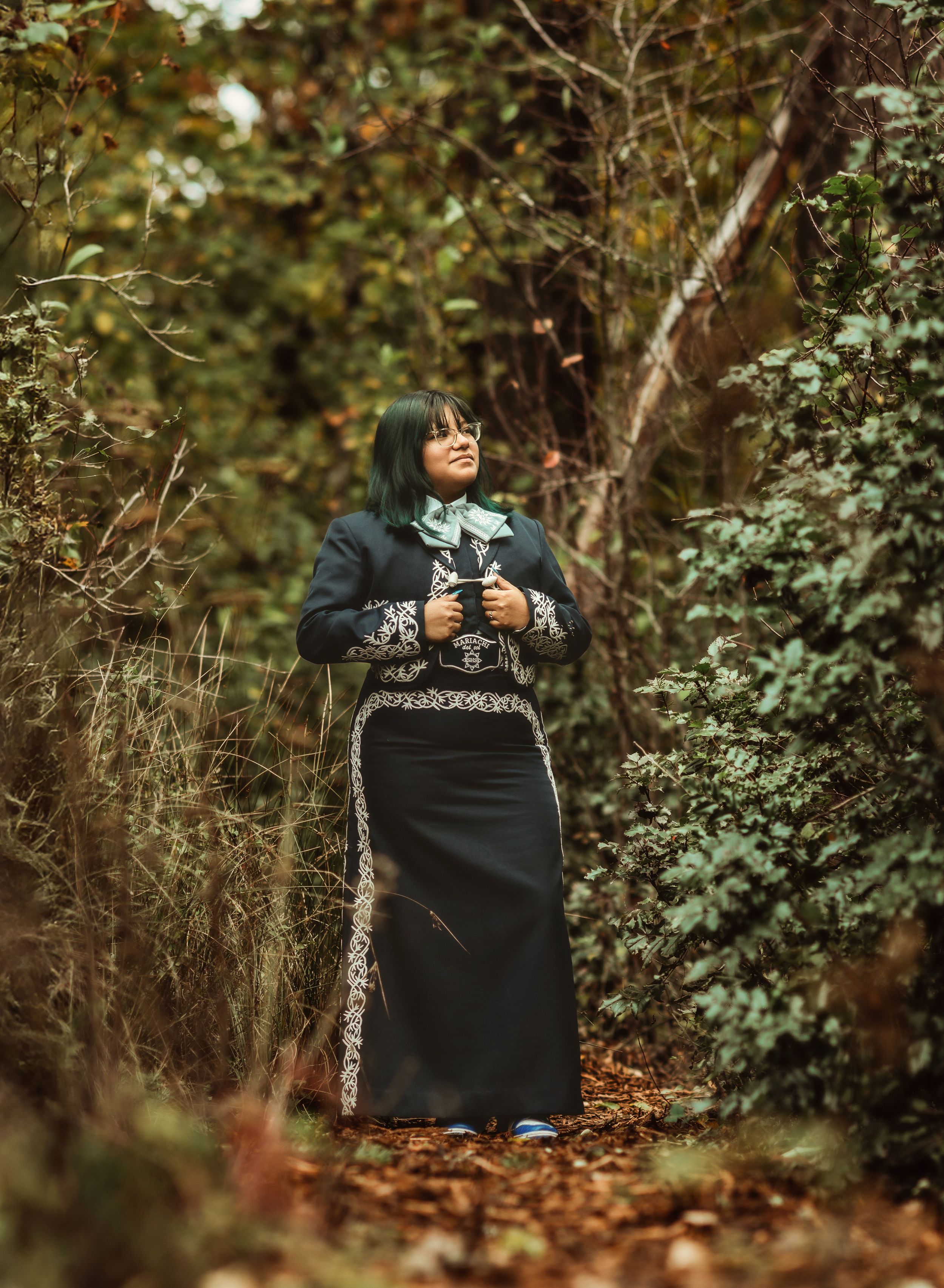 A person in a patterned sweater and long skirt stands on a woodland path holding a camera in autumn.