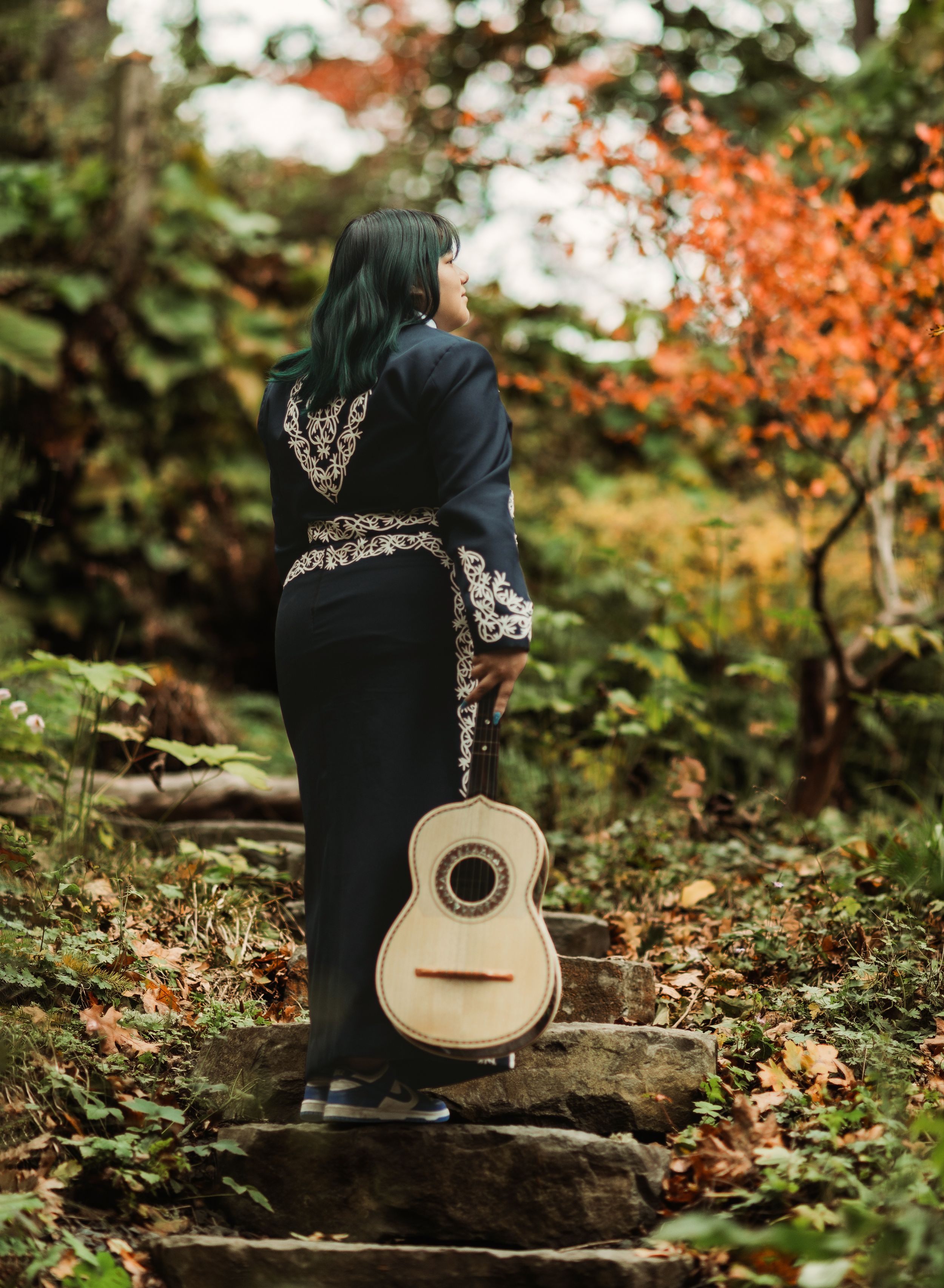 A person in a black jacket holds a guitar while sitting on stone steps surrounded by autumn foliage in a forest setting.