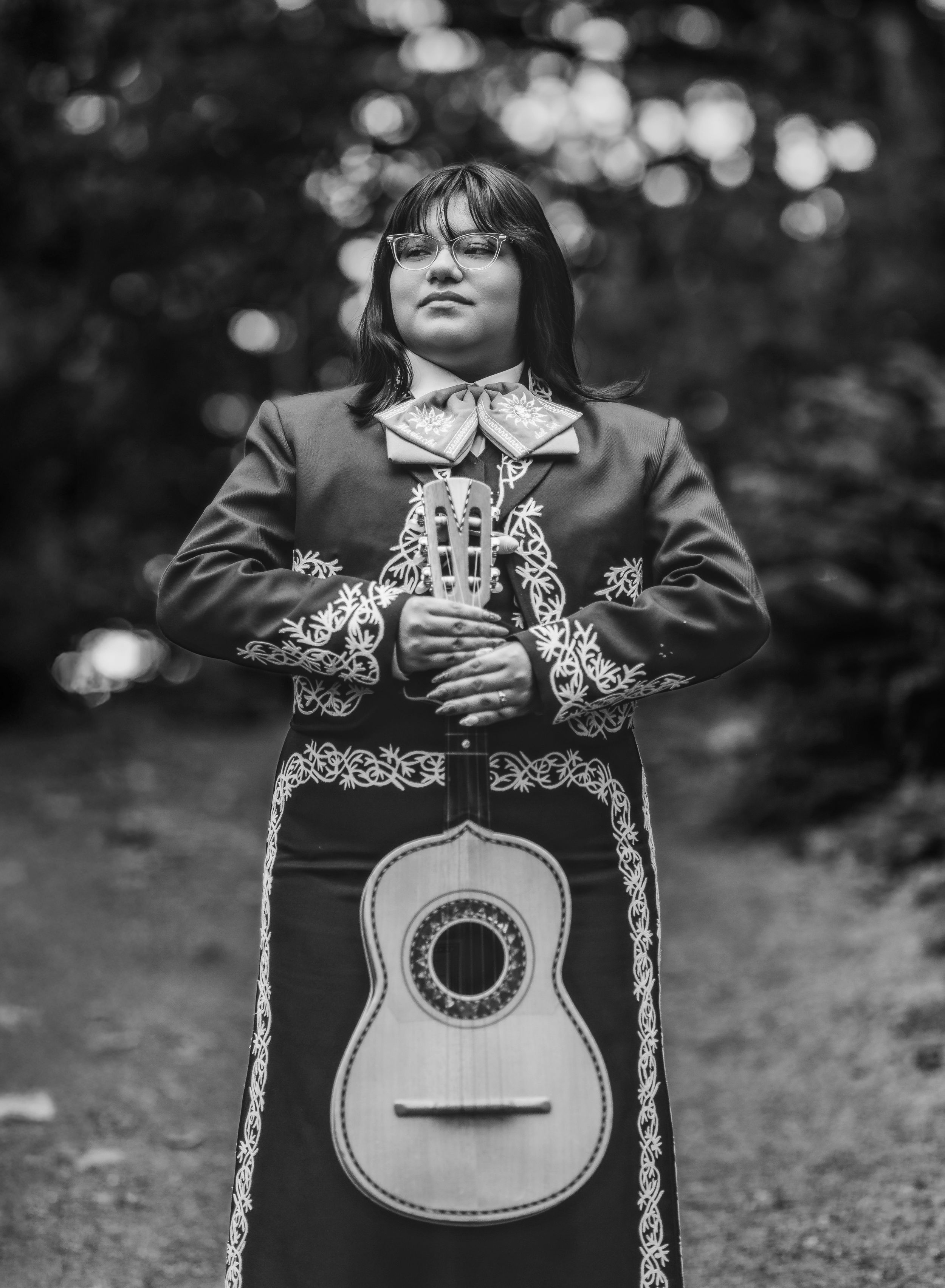 A mariachi musician in traditional black embroidered attire poses with their guitar in a black and white photograph.