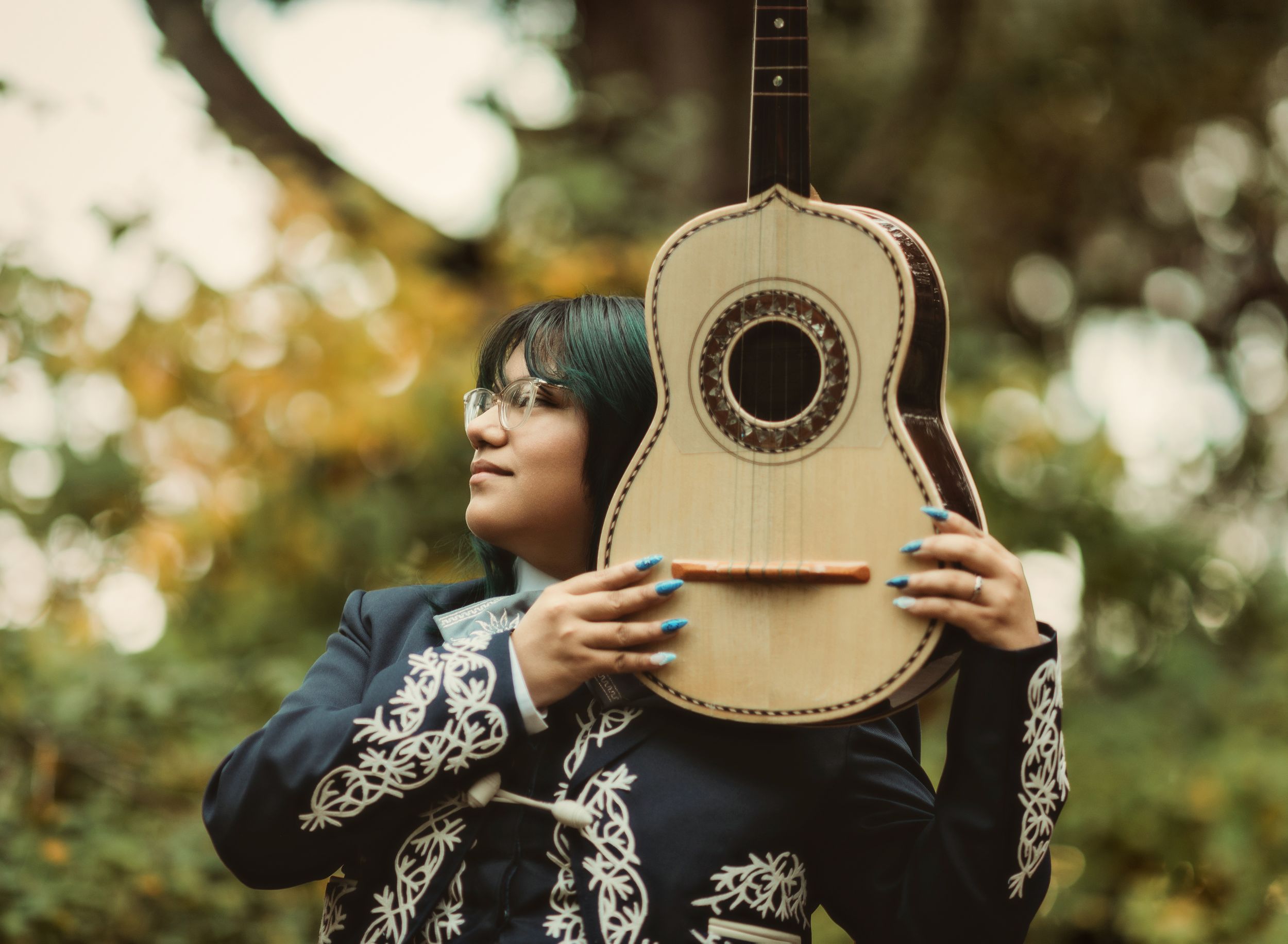 A person in a black and white embroidered dress holds a guitar outdoors in a sunlit garden setting.