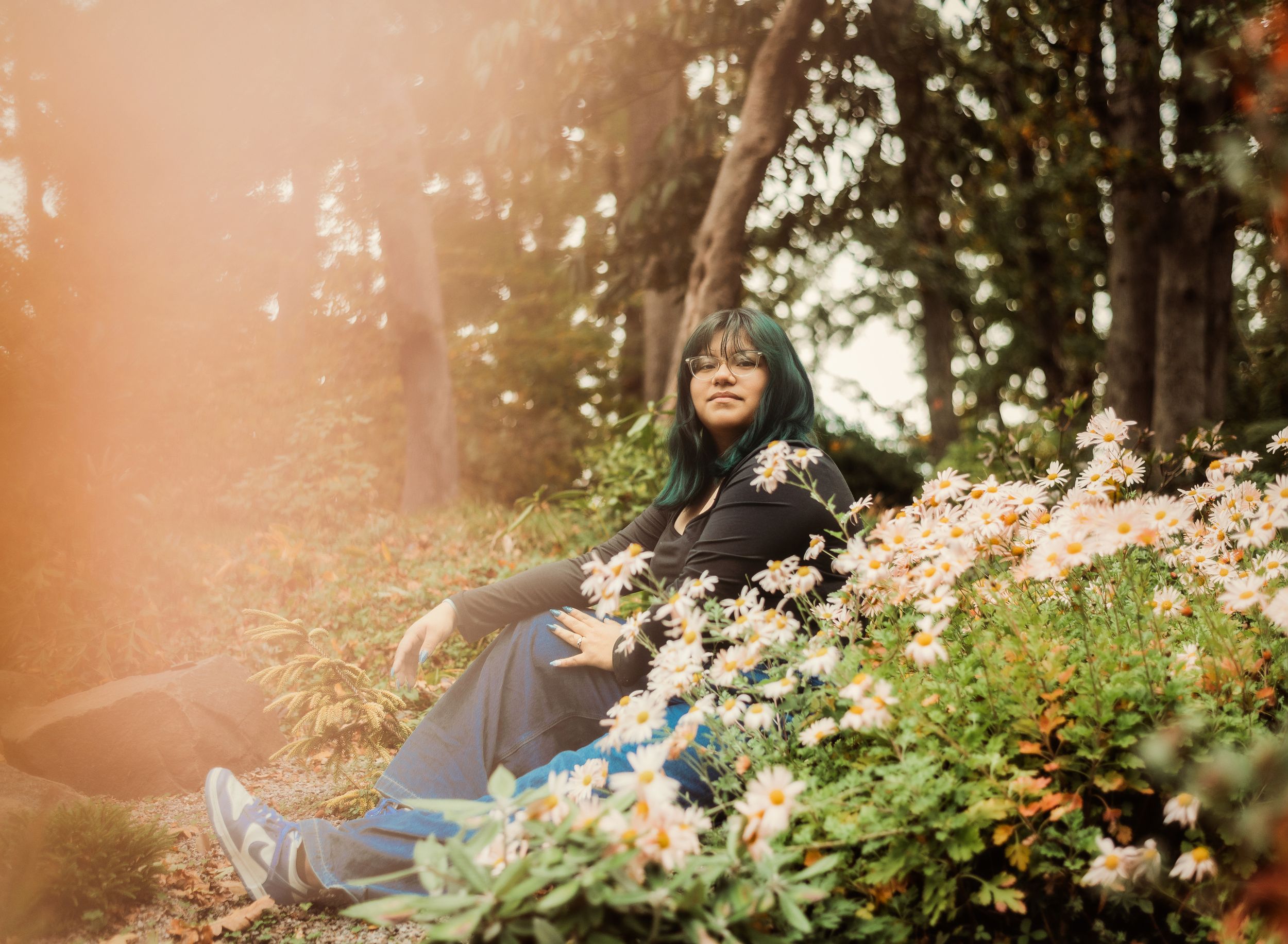 A person sits among blooming white flowers in a sunlit forest wearing casual attire and sneakers.