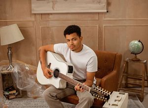 A person plays acoustic guitar while sitting in a vintage-style room with warm lighting and home decor elements.