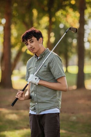 A young golfer in a gray shirt practices their swing form outdoors in a wooded park setting with golden sunlight.