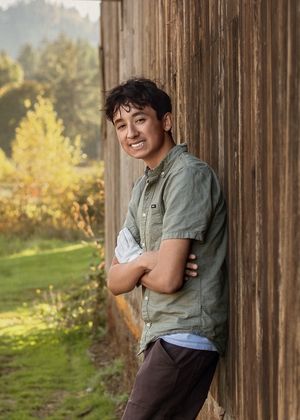 Person in casual olive shirt leans against rustic wooden wall with autumn foliage and sunlit meadow in background.