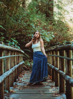 Young woman in blue maxi skirt and white top poses on a wooden bridge surrounded by lush green forest.