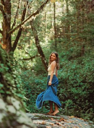 Woman in blue maxi skirt and cream top twirling barefoot on rocks in a lush green forest.