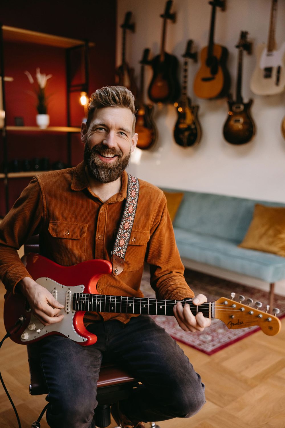 Musician in brown jacket playing red Fender Stratocaster in studio with guitars displayed on wall.