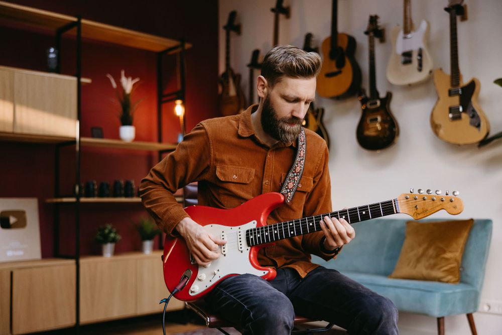 Musician playing red electric guitar in studio space with guitars displayed on wall.