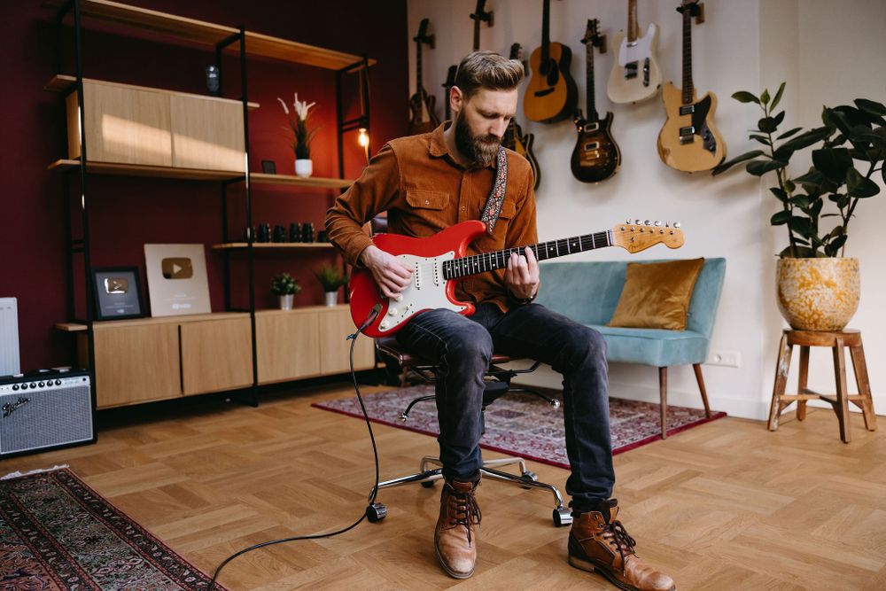 Musician playing red electric guitar in cozy studio space with wooden floors and guitar collection.