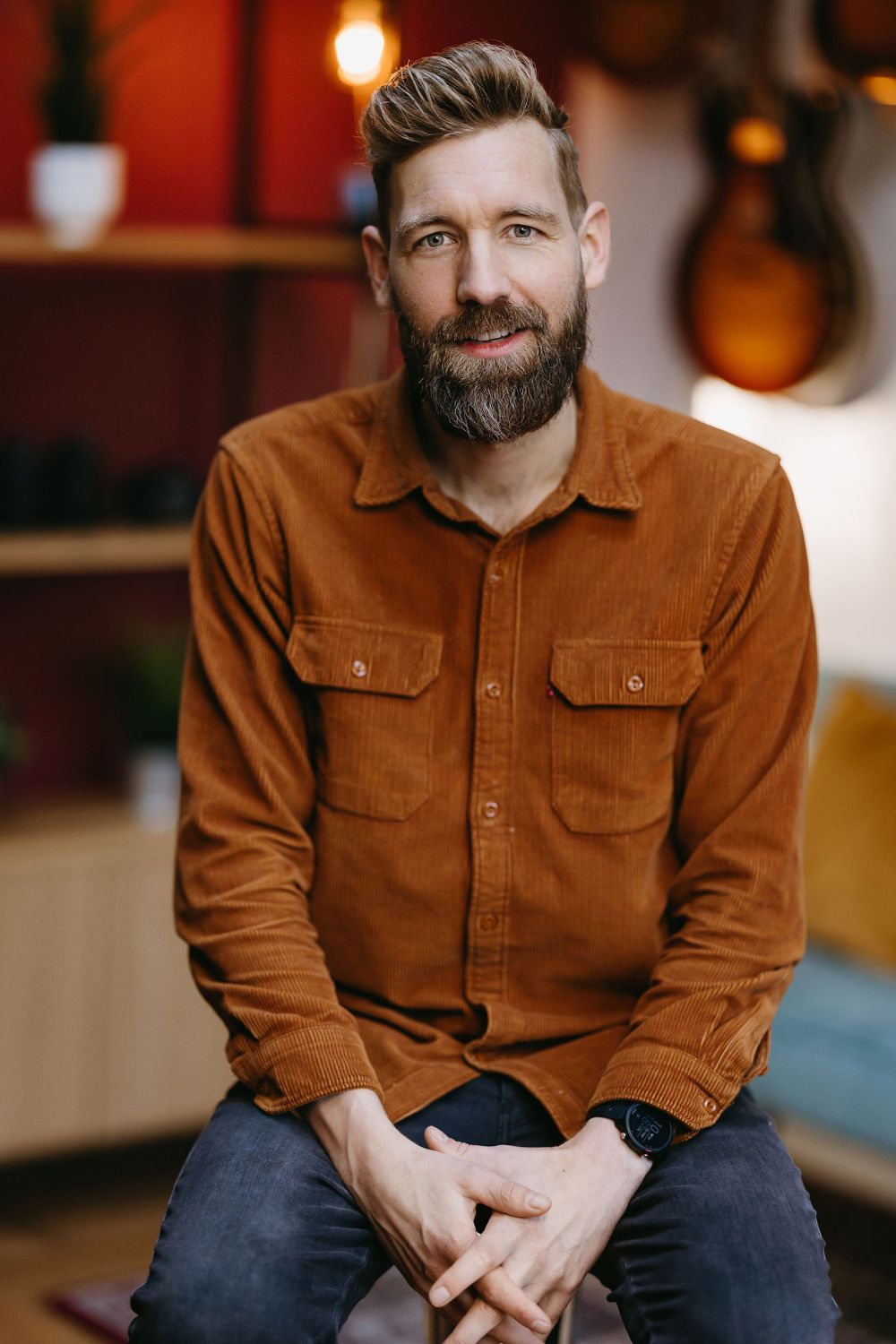 Professional musician in brown corduroy shirt sitting in front of guitar display wall.